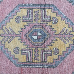 K&ouml;nnte beinhalten: Ein Vintage-Teppich mit einem geometrischen Muster in den Farben Rosa, Gelb, Blau und Grau. Der Teppich hat eine zentrale Diamantform mit einem kleineren Diamanten im Inneren. Der Teppich ist abgenutzt und verblasst, was ihm einen rustikalen Look verleiht.