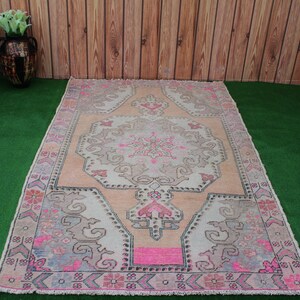 May include: A vintage-style rug with a pink and cream color scheme. The rug has a floral pattern with a central medallion. The rug is laid out on a green artificial grass surface.