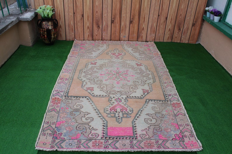 May include: A vintage-style rug with a pink and cream color scheme. The rug has a floral pattern with a central medallion. The rug is laid out on a green artificial grass surface.