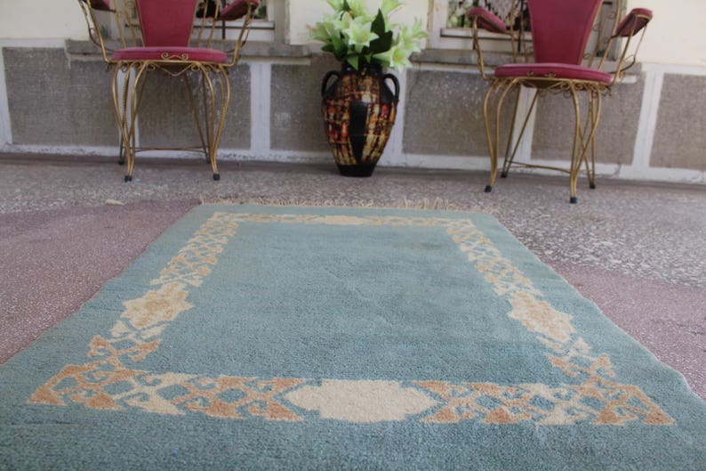 Op de afbeelding: Een lichtblauw tapijt met een beige gepatroneerde rand. Het tapijt ligt op een betonnen vloer voor twee stoelen met rode kussens.