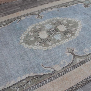 May include: A vintage rug with a light blue background and a central medallion in shades of green and white. The rug has a border with a repeating pattern of small white and brown shapes.
