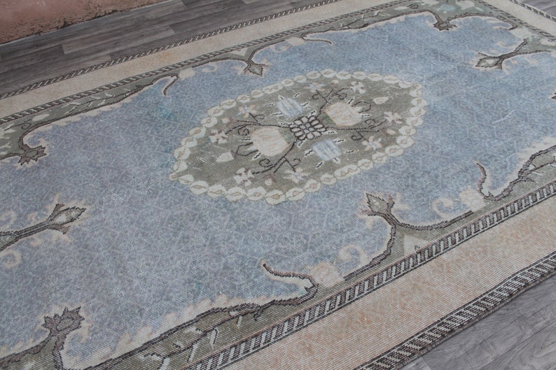 May include: A vintage rug with a light blue background and a central medallion in shades of green and white. The rug has a border with a repeating pattern of small white and brown shapes.