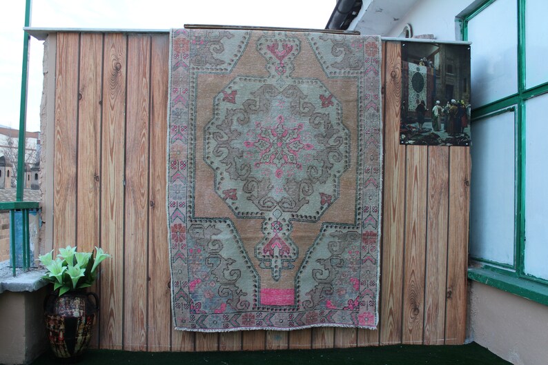 May include: A vintage rug with a light pink, beige, and gray color scheme. The rug has a traditional Persian design with a central medallion and intricate floral patterns. The rug is hanging on a wooden wall.