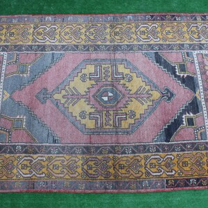 K&ouml;nnte beinhalten: Ein Vintage-Teppich mit einem geometrischen Muster in Rosa, Blau und Gelb. Der Teppich hat einen dunkelbraunen Rand mit komplizierten Designs.