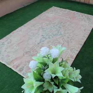 Puede incluir: Una alfombra vintage de color rosa claro con un patr&oacute;n floral, colocada sobre una superficie de c&eacute;sped artificial verde. Un ramo de flores artificiales blancas y verdes se encuentra en primer plano.