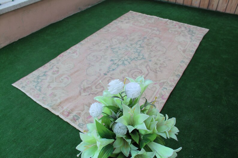 Puede incluir: Una alfombra vintage de color rosa claro con un patr&oacute;n floral, colocada sobre una superficie de c&eacute;sped artificial verde. Un ramo de flores artificiales blancas y verdes se encuentra en primer plano.