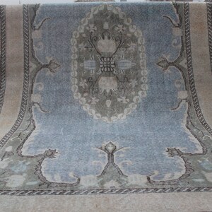 May include: A vintage rug with a light blue background and a central medallion in shades of green, white, and beige. The rug has a cream-colored border with a repeating pattern.