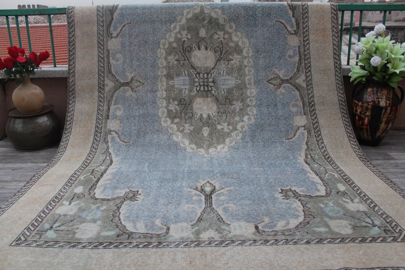 May include: A vintage rug with a light blue background and a central medallion in shades of green, white, and beige. The rug has a cream-colored border with a repeating pattern.