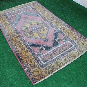 K&ouml;nnte beinhalten: Ein vintage t&uuml;rkischer Teppich mit einem geometrischen Muster in Rosa, Gelb und Blau. Der Teppich hat einen Fransenrand und liegt auf einer gr&uuml;nen Oberfl&auml;che.