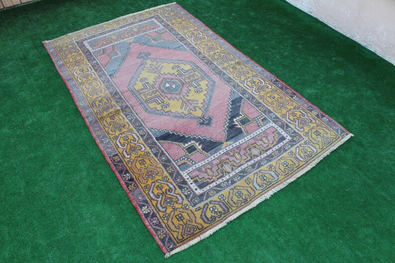 K&ouml;nnte beinhalten: Ein vintage t&uuml;rkischer Teppich mit einem geometrischen Muster in Rosa, Gelb und Blau. Der Teppich hat einen Fransenrand und liegt auf einer gr&uuml;nen Oberfl&auml;che.
