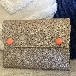Peut inclure: Une petite pochette dorée à paillettes avec un rabat et deux boutons circulaires orange. La pochette est rectangulaire et semble être faite d'un matériau texturé. L'arrière-plan comprend un oreiller blanc et un oreiller bleu.