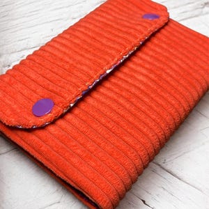 Op de afbeelding: Een oranje corduroy portemonnee met een paarse knoopsluiting. De portemonnee heeft een getextureerd, geribbeld oppervlak en een voering met patroon. De ronde knoop zorgt voor een veilige sluiting.