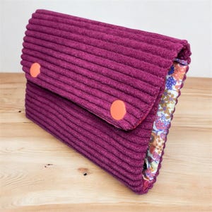 Op de afbeelding: Een pruimkleurige corduroy clutch met twee oranje ronde knopen. De binnenvoering heeft een bloemenpatroon met tinten blauw, roze en geel. De clutch is gesloten en ligt op een licht houten oppervlak.