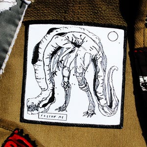 Pode incluir: Close-up de uma bolsa de lona com vários patches. Os patches incluem um desenho a preto e branco de um coelho, uma criatura com o texto "FOLLOW ME", uma foto policial e um patch com a palavra "CRUCIFY". A bolsa é bege.