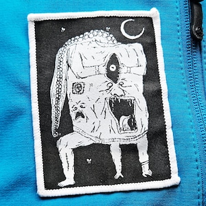 Puede incluir: Un parche en blanco y negro con un diseño de monstruo y una luna creciente. El parche rectangular con borde blanco está adherido a una tela azul turquesa. El monstruo tiene un ojo y una boca abierta.
