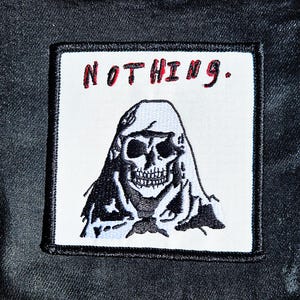Pode incluir: Patch bordado a preto e branco com uma caveira com capuz e a palavra "NOTHING." em vermelho. Costurado numa jaqueta jeans preta.