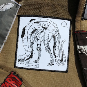 Peut inclure: Une veste en toile marron avec différentes patchs cousues. Les patchs présentent une illustration en noir et blanc d'un lapin, une illustration en noir et blanc d'une créature à plusieurs membres, une illustration en noir et blanc d'une personne pendue à une croix et une illustration en noir et blanc d'une personne tenant une pancarte qui dit "Hollywood Police Dept.  Battered"