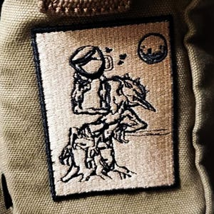 May include: A tan canvas bag with a rectangular patch showing a black and tan illustration of a creature with a bird-like head and a person holding a mug. A small circle with a cityscape is in the upper right corner.