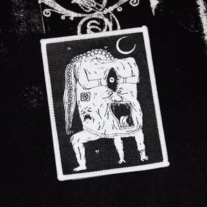 Puede incluir: Un parche bordado rectangular en blanco y negro que presenta una ilustración surrealista de una criatura con un gran ojo central, una boca muy abierta y un largo apéndice similar a un tentáculo sobre un fondo oscuro con una luna creciente.