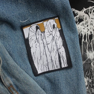 Puede incluir: Parche bordado en blanco y negro de dos figuras con túnicas con capucha, sobre un fondo dorado. El parche está cosido en una chaqueta vaquera azul.