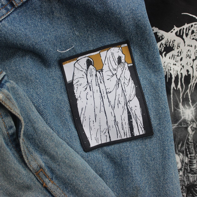 May include: A rectangular patch featuring a black and white illustration of two figures draped in white cloaks, with a gold background. The patch is displayed on a blue denim jacket, with a glimpse of a black graphic tee in the background.