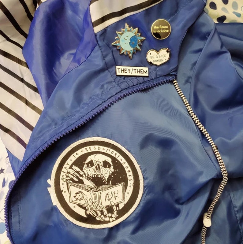 May include: A blue windbreaker jacket featuring a circular patch with a skeleton reading a book and the text "TRY TO READ MORE BOOKS". Several enamel pins are attached, including a sun and moon, a heart reading "BE A NICE HUMAN", "THEY/THEM", and a round pin stating "the future is inclusive".