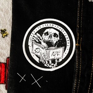 May include: A circular black and white embroidered patch featuring a skeleton holding an open book. The text "TRY TO READ MORE BOOKS" is printed along the upper border. The patch is attached to a black fabric surface with visible stitching.