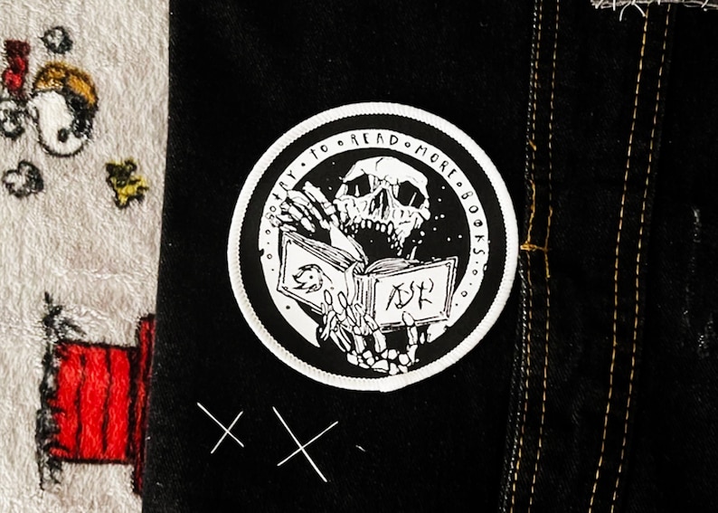 May include: A circular black and white embroidered patch featuring a skeleton holding an open book. The text "TRY TO READ MORE BOOKS" is printed along the upper border. The patch is attached to a black fabric surface with visible stitching.