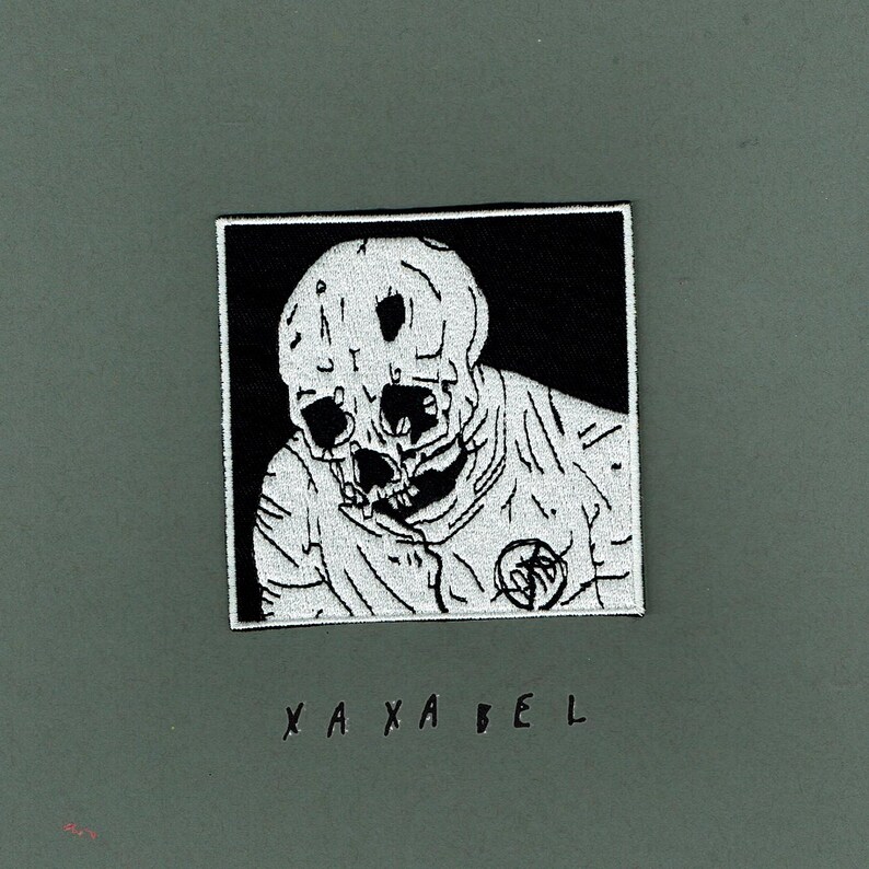 May include: A square embroidered patch featuring a white skull and upper torso graphic on a black background. The design includes a circular emblem on the chest area. Below the patch, the text XA XA BEL is printed in black on the surface.