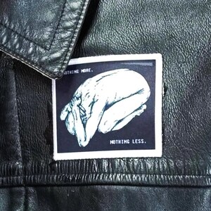 May include: A black leather jacket featuring a rectangular patch with a white border. The patch displays a dark blue background with a white illustration of a person in a fetal position, accompanied by the text NOTHING MORE. and NOTHING LESS.