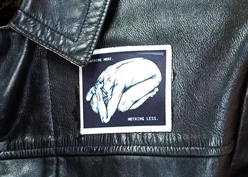May include: A black leather jacket featuring a rectangular patch with a white border. The patch displays a dark blue background with a white illustration of a person in a fetal position, accompanied by the text NOTHING MORE. and NOTHING LESS.