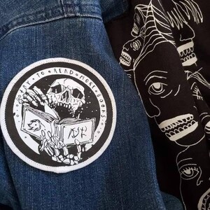 May include: A circular black and white embroidered patch featuring a skeleton reading a book with the text "TRY TO READ MORE BOOKS" along the border, attached to a blue denim fabric next to a black textile with a white line art face pattern.