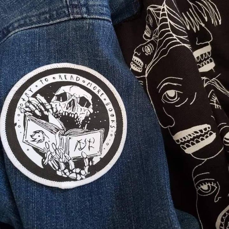 May include: A circular black and white embroidered patch featuring a skeleton reading a book with the text "TRY TO READ MORE BOOKS" along the border, attached to a blue denim fabric next to a black textile with a white line art face pattern.