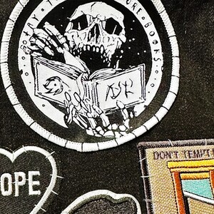 May include: A black denim fabric featuring three embroidered patches. A large circular patch shows a skeleton reading a book with the text "SO MANY BOOKS". A heart-shaped patch reads "NOPE". A rectangular patch depicts a guillotine with "DON'T TEMPT ME".