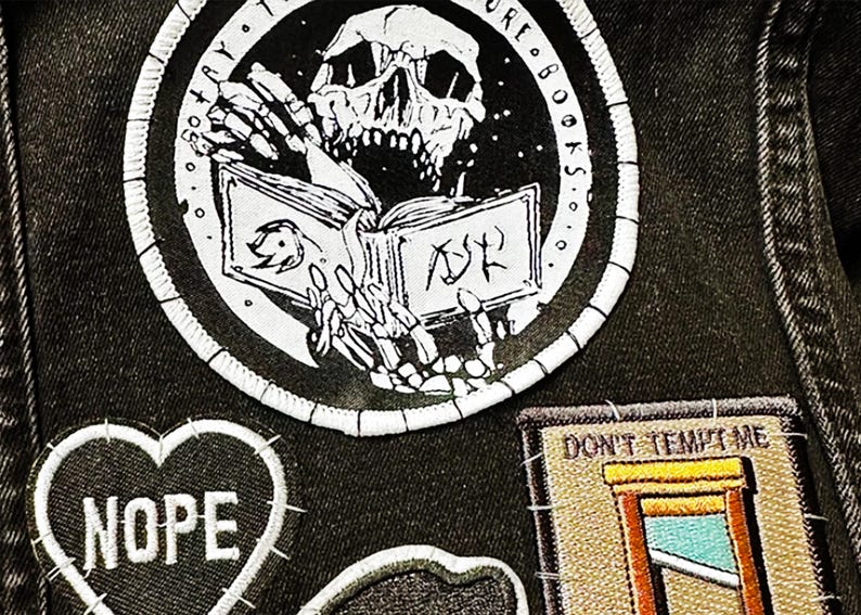 May include: A black denim fabric featuring three embroidered patches. A large circular patch shows a skeleton reading a book with the text "SO MANY BOOKS". A heart-shaped patch reads "NOPE". A rectangular patch depicts a guillotine with "DON'T TEMPT ME".