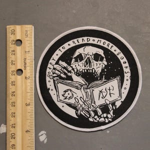 May include: A circular black and white embroidered patch featuring a skeleton reading an open book. The text "TRY TO READ MORE BOOKS" is arched along the top border. A wooden ruler is placed to the left, showing the patch is approximately 3 inches wide.