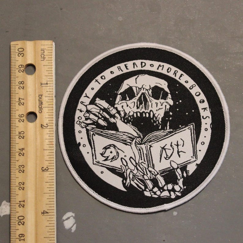 May include: A circular black and white embroidered patch featuring a skeleton reading an open book. The text "TRY TO READ MORE BOOKS" is arched along the top border. A wooden ruler is placed to the left, showing the patch is approximately 3 inches wide.