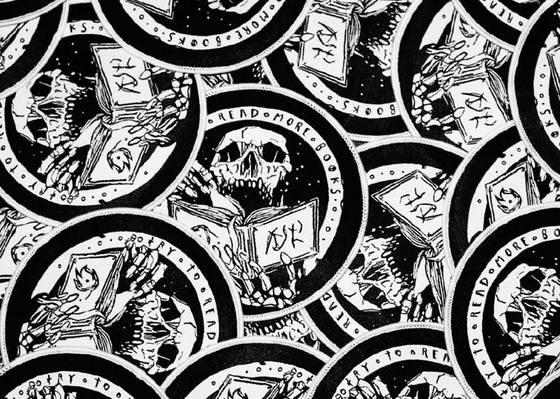 May include: A collection of circular black and white embroidered patches featuring a skeletal figure holding an open book. The text READ MORE BOOKS and TRY TO READ is printed along the circular border of each patch, surrounding the central skull.