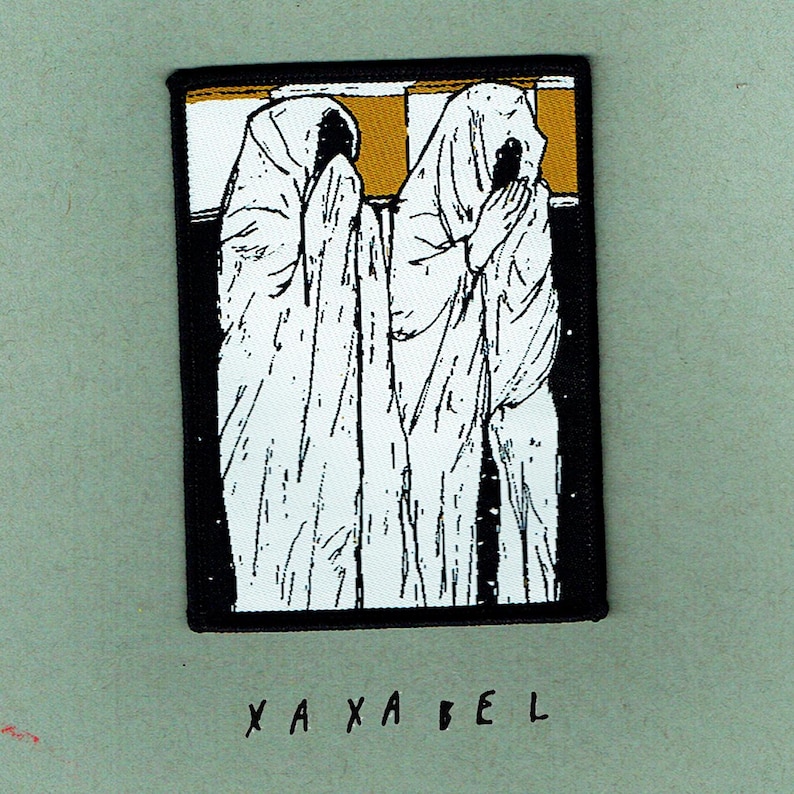 May include: A rectangular patch featuring a black border and a design of two figures in white robes with obscured faces. The background includes gold and white stripes. The word "XAXABEL" is written in black at the bottom.