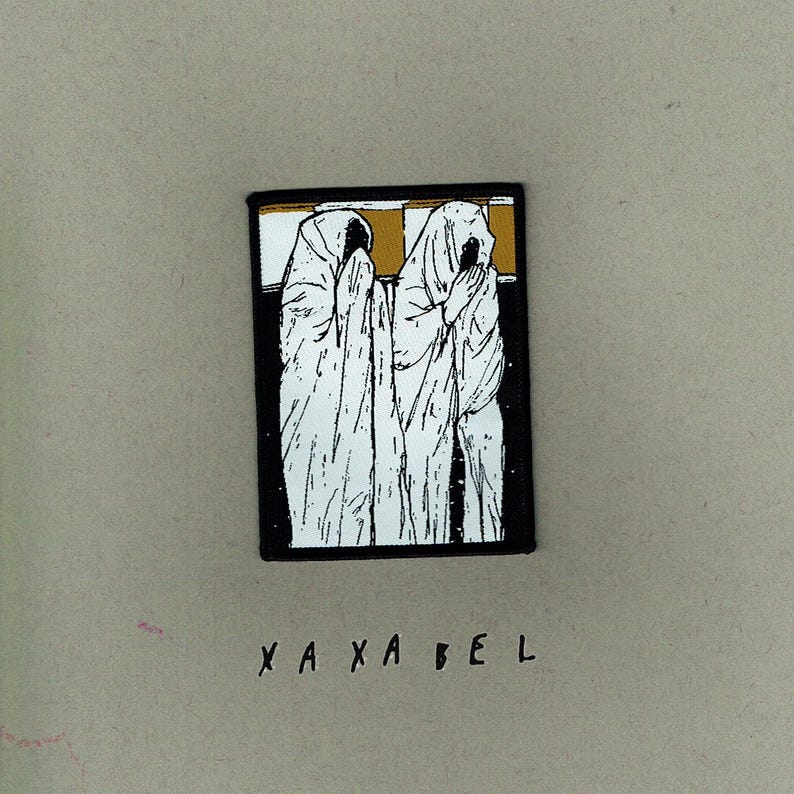 May include: A black patch featuring a white illustration of two figures in hooded robes, with the text "X A X A B E L" in black at the bottom. The patch has a gold and black border.