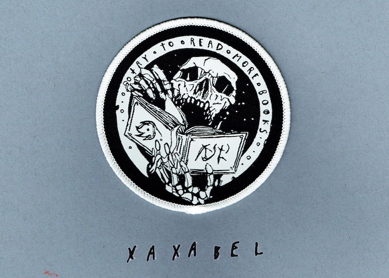 May include: A circular embroidered patch with a white border featuring a black and white illustration of a skeleton reading an open book. The text "TRY TO READ MORE BOOKS" is printed in a circular arc along the top edge of the black background.