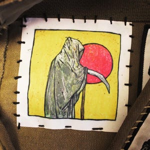 May include: A hand-stitched patch depicting the Grim Reaper with a scythe, set against a yellow backdrop and a red sun. The patch is framed in white and sewn onto a canvas material.