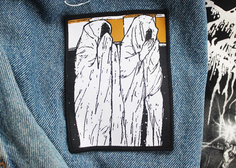May include: A rectangular patch featuring a black and white illustration of two figures draped in white cloth, with hands clasped in prayer. The background is a mix of gold and black. The patch is sewn onto a blue denim jacket.