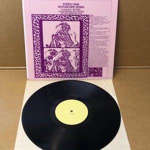 Può includere: Un album in vinile vintage di Steely Dan, intitolato "Rotoscope Down". La copertina dell'album è rosa con testo e grafica viola. Il disco è nero con un'etichetta centrale gialla. Il testo include i titoli delle canzoni.
