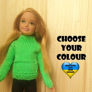 May include: A doll wearing a green knitted sweater and black trousers. The doll has long blonde hair and blue eyes. The background is a wooden surface with the text "Choose your colour" and "Made with love from Ukraine".