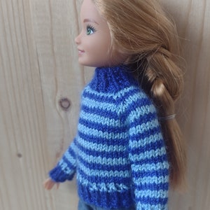 May include: A doll wearing a blue and light blue striped sweater with a turtleneck. The doll has long blonde hair and is wearing blue jeans.
