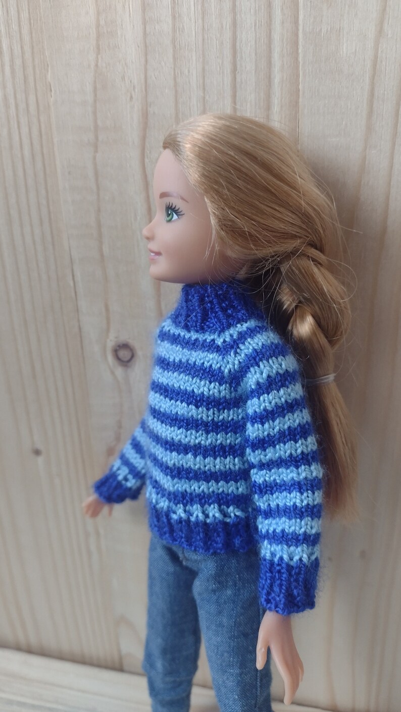 May include: A doll wearing a blue and light blue striped sweater with a turtleneck. The doll has long blonde hair and is wearing blue jeans.