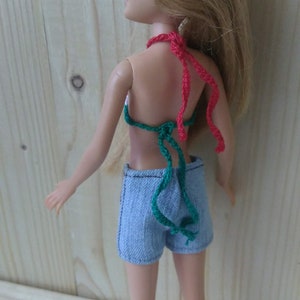 May include: A doll wearing a green and red crocheted bikini top and blue denim shorts. The doll has long blonde hair and is standing in front of a wooden background.