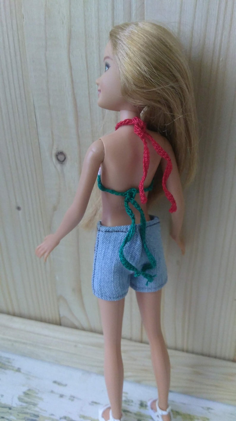 May include: A doll wearing a green and red crocheted bikini top and blue denim shorts. The doll has long blonde hair and is standing in front of a wooden background.