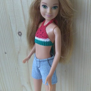 May include: A doll with long blonde hair wearing a red, green and white watermelon-patterned halter top, blue denim shorts and white sandals.